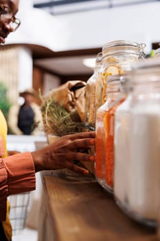 In local store, African American woman looks for fresh, organic veggies. A close-up view of a hand touching glass containers filled with spaghetti. Reusable packaging, sustainable choices.