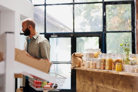 Middle Eastern man browsing different food items in other sections of the store. Male consumer holds basket filled with a mixture of various fruits and vegetables, healthy blend of bio products.