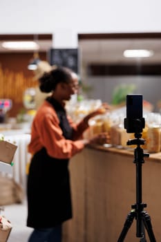 Black woman at local supermarket, promoting healthy and sustainable living. A smartphone is used to shoot a vlog to highlight eco friendly solutions, fresh vegetables and glass jars. Foreground focus.