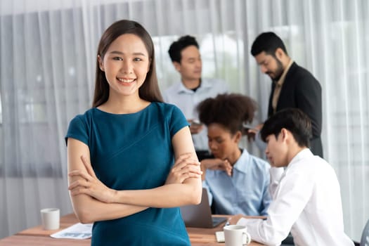 Young Asian businesswoman poses confidently with diverse coworkers in busy meeting room background. Multicultural team works together for business success. Office lady portrait. Concord