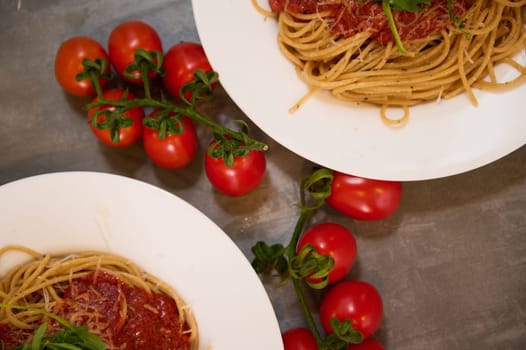 Top Partial view of two white plates with Italian freshly cooked capellini pasta with delicious tomato sauce, garnished with green leaves of arugula, near a bunch of cherry tomatoes. Food background