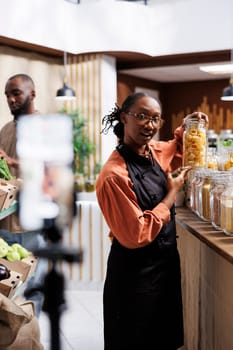 Young black woman records a video in a zero-waste shop promoting fresh, organic products. She uses a tripod stand while showcasing sustainable packaging and healthy options.