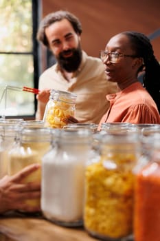 Portrait of african american woman holding a glass jar filled with pasta while standing next to caucasian man in local store. Multicultural couple doing sustainable shopping for healthy lifestyle.