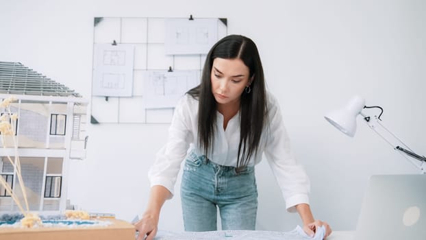Young beautiful caucasian architect puts blueprint on the table and compare with house model while inspect hos model construction carefully at modern office with blueprint striped behind. Immaculate.