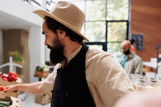 Portrait shot of vendor with hat and apron looking and holding carrots in a box on shelf in an eco friendly store. Caucasian salesman vlogging about various fresh produce in his sustainable market.