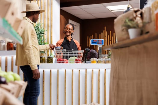 A young African American man in a hat shops in a contemporary supermarket, browsing items and paying with his credit card. The POS terminal at the checkout counter is operated by the shop owner.