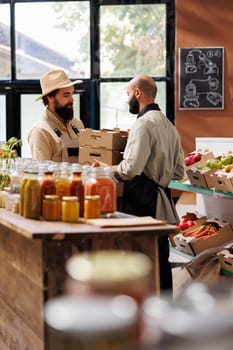 Caucasian man handing over boxes of freshly harvested items to middle eastern vendor with black apron in grocery store. Storekeeper receives new batch of locally grown produce from farmer.