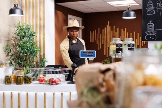 Focus on background of african american male shopkeeper at the checkout counter. Black man wearing a hat, arranging items over at the cashier desk in bio-store. Fresh, eco friendly produce displayed.