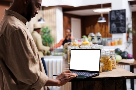 An eco friendly supermarket uses a digital device to display its products. African American male customer with a laptop showing an isolated white screen is browsing for bio-food items.