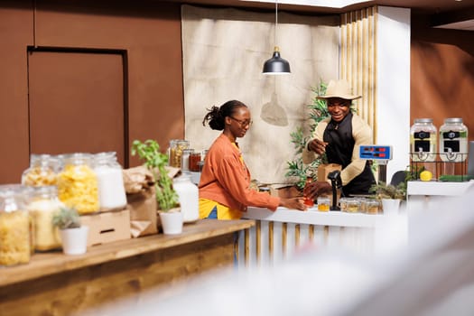 In grocery store, an African American shop owner advises an environmentally conscious client with fresh organic groceries. Male vendor encourages a zero-waste lifestyle by providing reusable bags.