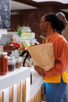 Image of black woman with a grocery bag handing over red pepper to the African American local vendor. A smiling female customer giving her selected fresh produce to the cashier, sustainable shopping.