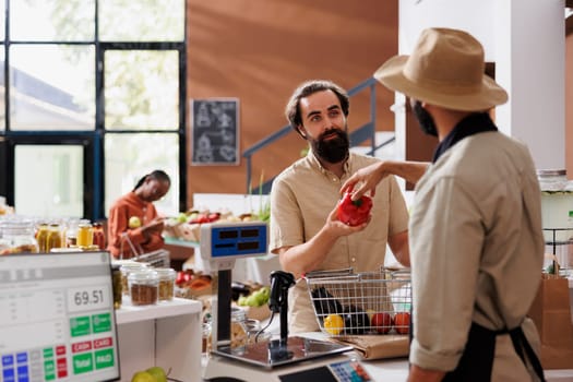 A young salesman wearing an apron and hat is helping caucasian customer buy fresh, environmentally friendly foods in a nearby store. Various nutritious food options and recyclable packaging.