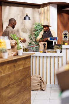 At the cashier desk, an African American shopkeeper serves a customer. A guy seller wearing an apron places products in a brown paper bag for the black man at the counter. Background focus.