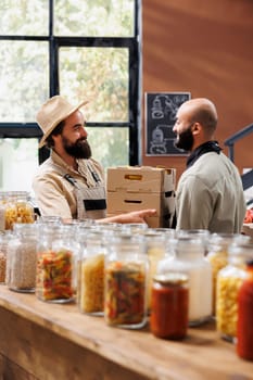 Smiling man delivering handpicked eco friendly food products to middle eastern salesman in black apron. Caucasian farmer handing crates full of healthy fruits and vegetables to seller.