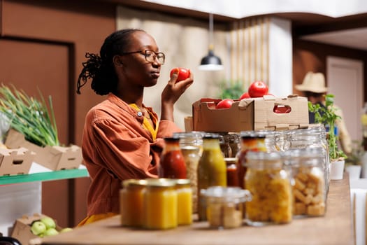 Black woman shops at local market, supporting local producers and choosing eco friendly products like fresh fruits and vegetables. African American lady near the glass checking out the tomatoes.
