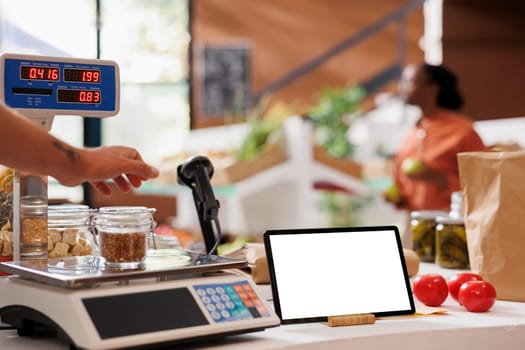 Glass jar filled with bio food items is placed on a measuring weight scale on the counter of grocery store. Image shows a tablet displaying blank chromakey mockup template while seller weighs an item.