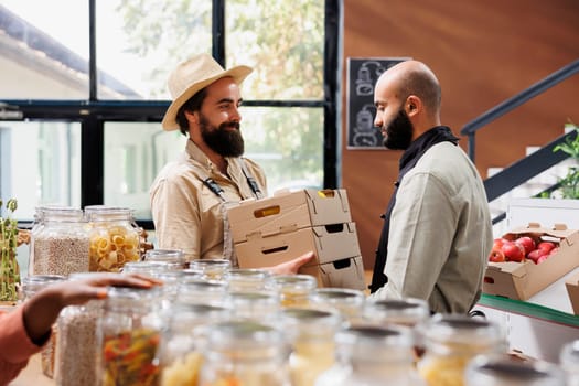 Farmer providing locally sourced vegetables to middle eastern vendor selling them to healthy living customers in bio shop. Man handing crates full of additives free fresh produce to retailer.