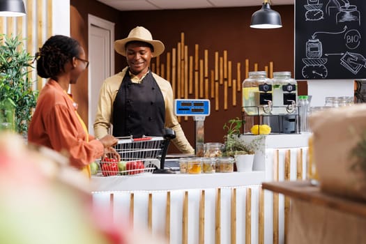 Friendly black male cashier with warm smile assists female client with her purchase at local market checkout counter. The image showcases excellent customer service and diverse shopping experience.