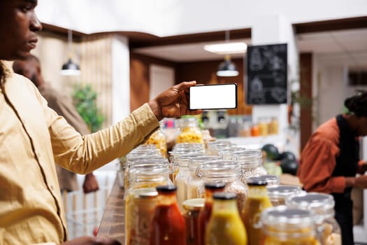 At eco friendly grocery shop African American male consumer holds a cell phone that displays an isolated white screen. Image shows black man with mobile device having a chromakey template.