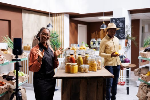 African American business owner films vlogs promoting fresh, organic products in the eco friendly store, using smartphone and selfie stick. Image shows black woman speaking in front of mobile device.