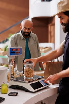 Salesman wearing an apron and using digital weighing scale to measure glass jar filled with organic soybeans. Male customer waiting while caucasian vendor checks price of bulk product.