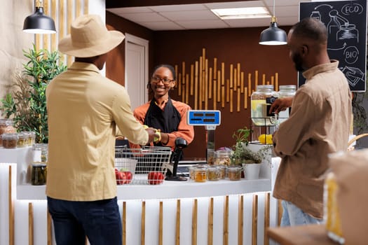 In grocery store, African American female vendor assists clients, promoting sustainability with fresh products in bio packaging. At counter, black woman taking items from male customer for weighing.