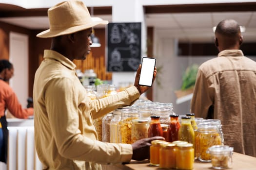 African American male customer holding mobile device displaying blank copyspace chromakey template at an eco friendly grocery store. Black man grasping a smartphone with white screen.