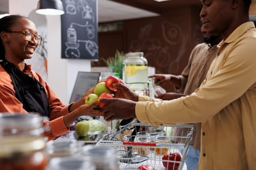 African American owner helps customers at eco friendly store with fresh organic products. Selective focus on a male client handing fruits to the female cashier at the checkout counter. side view shot.