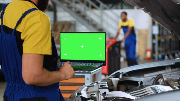 Licensed repairman using green screen laptop in car service to order new parts for busted vehicle. Close up shot of professional using chroma key device to look on internet for replacing components