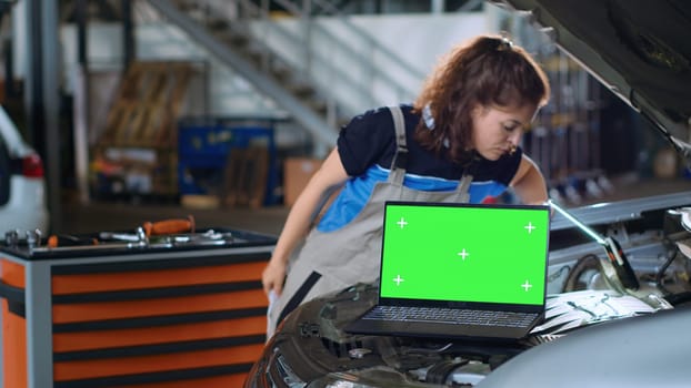 Chroma key laptop in garage workplace sitting on opened up car. Mockup device in auto repair shop next to mechanic cleaning vehicle inside engine compartment after fixing it