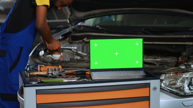 Close up shot of laptop placed on working bench in busy garage next to professional tools while serviceman fixes vehicle. Mockup device in auto repair shop with mechanic working in blurry background