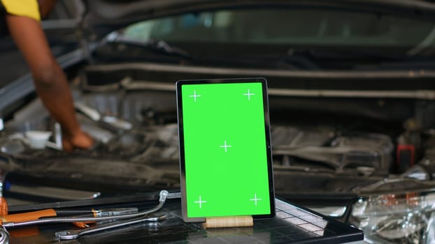 Close up shot of tablet placed on working bench in busy garage next to professional tools while engineer fixes vehicle. Mockup device in repair shop with precise expert working in blurry background