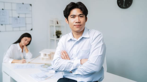 Professional asian businessman crosses arms with confident while young beautiful caucasian colleague drawing a blueprint on meeting table with house model and blueprint at modern office. Immaculate.