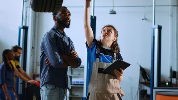 Mechanic in garage stands with customer underneath car on overhead lift, looking together for engine replacement. Expert helps BIPOC client recondition vehicle using tablet to find needed part