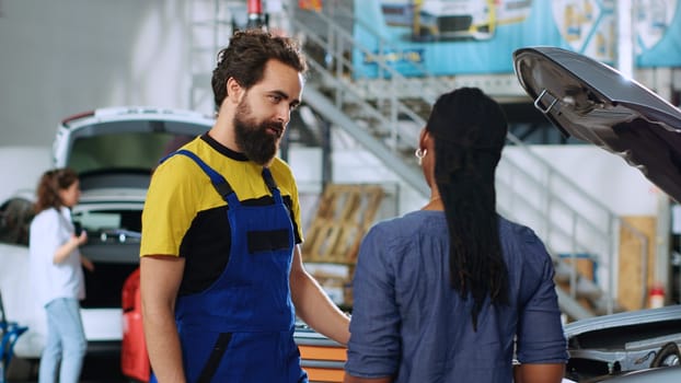 Engineer in auto repair shop showing client malfunctions found inside car after thorough inspection. Repairman telling african american woman vehicle diagnostic after checking for broken parts