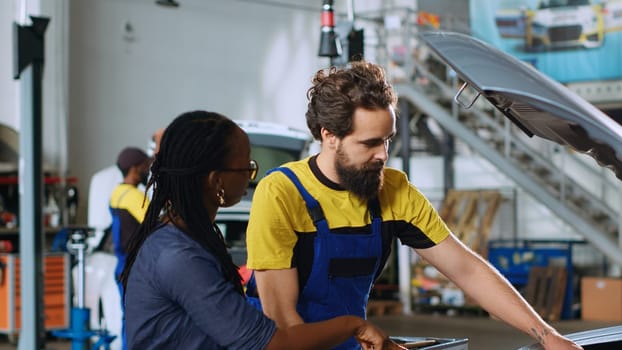 Qualified serviceman in repair shop showing client damages found inside car after inspection. Automobile specialist telling woman vehicle diagnostic after checking for defective parts