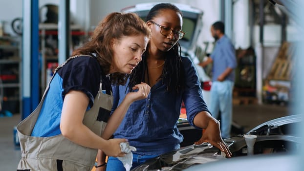 Technician in garage workspace cleaning opened up vehicle after replacing brake fluid while customer watches. Car service worker wiping automobile leftover leakage and discussing with woman