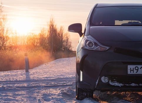 A dark gray car is parked on a snow-covered road as the sun rises in a winter countryside setting, creating a serene and peaceful atmosphere.