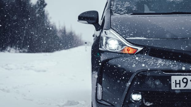 A dark gray car is parked on a snow-covered road at snowfall in a winter countryside setting, creating a serene and peaceful atmosphere.