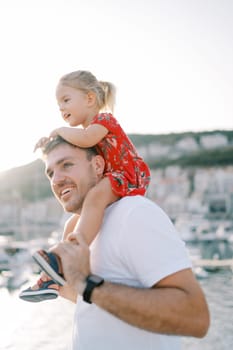 Dad with a smiling little girl on his shoulders walks along the seashore and looks to the side. High quality photo