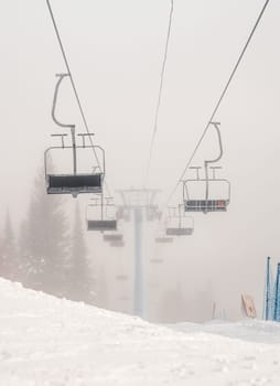 Ski lifts hang empty on a foggy morning at a snow-covered mountain resort, surrounded by frosty trees and blanketed in mist, creating a serene and quiet atmosphere.