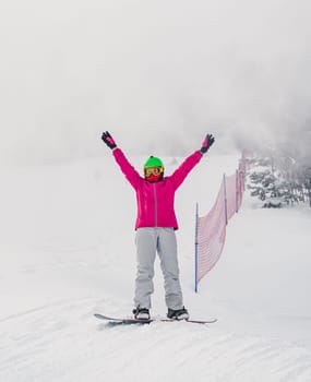 A snowboarder celebrates with arms raised on a misty, snow-covered slope. The scene is set in a winter landscape with some trees and a safety net in the background.