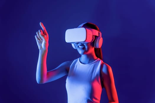 Smart female stand with surrounded by cyberpunk neon light wear VR headset connecting metaverse, futuristic cyberspace community technology. Woman using finger pointing virtual object. Hallucination.
