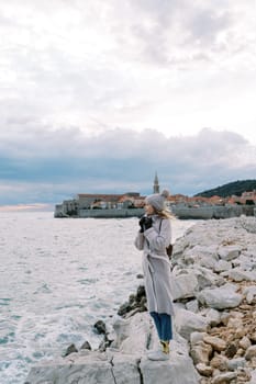 Young woman stands on a rocky shore wrapped in a coat against the background of the coast of an old town. High quality photo