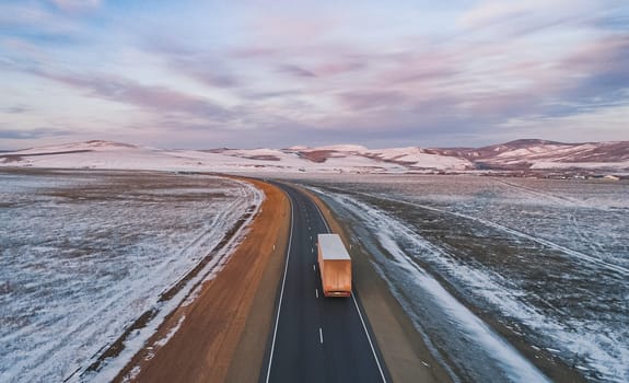 A lone semi truck drives along a winding rural highway through a snow-covered landscape during dusk in winter.