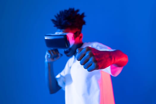 African American boxing gamer wearing VR playing boxer game punch camera posing isolated blue pink neon light wall connecting futuristic technology virtual reality metaverse fighting box. Contrivance.