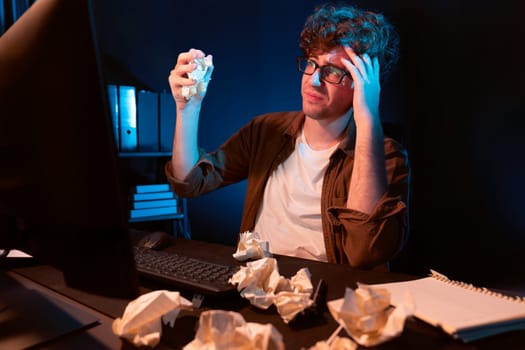 Stressful creative manager working with overworked project surrounded crumpled papers while waiting email sending back on laptop screen in casual shirt at night time at neon modern office. Gusher.
