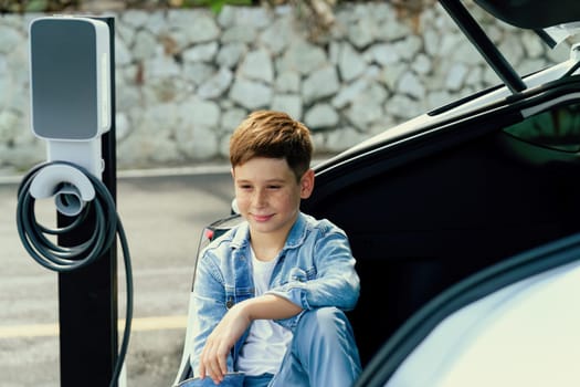 Little boy sitting on car trunk while recharging eco-friendly electric car from EV charging station. EV car road trip travel concept for alternative transportation powered sustainable energy.Perpetual