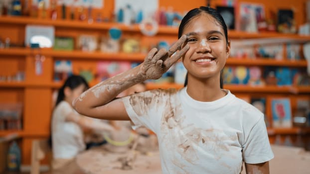 Happy caucasian girl pose at camera while diverse children modeling clay behind. Cute student wearing dirty shirt while looking at camera at workshop in art lesson. Blurring background. Edification.