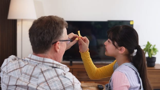 Grandfather and granddaughter eat french fries as food watch entertainment on TV. Old senior use technology communicate with young generation cross generation gap strengthen family bond. Divergence.